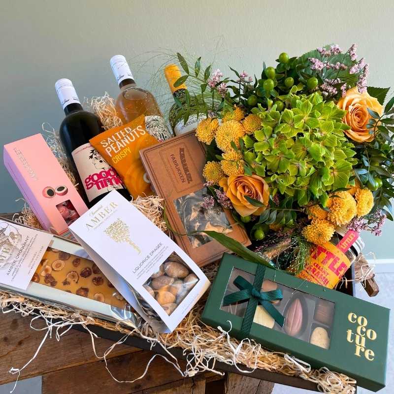 Gavekurv med 3 forskellige slags vin, forskellige chokolader og en buket i orange og grønne farver