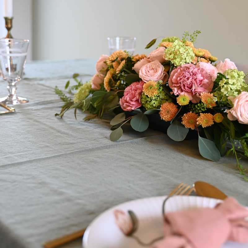 Borddekoration i aflang form med blomster i lyserøde, sart orange og lime farver på opdækket middagsbord med grøn dug