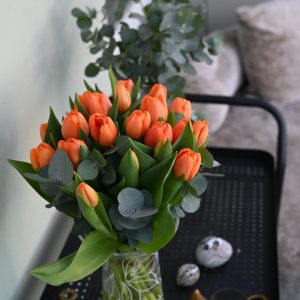 Buket orange tulipaner i en gennemsigtig glasvase, der står på et sort rullebord i hjemlige omgivelser.