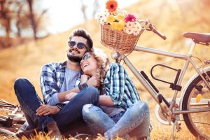 En mand og en kvinde sidder på jorden og griner tæt. Bagved dem står en cykel, hvor der er blomster i cykelkurven.