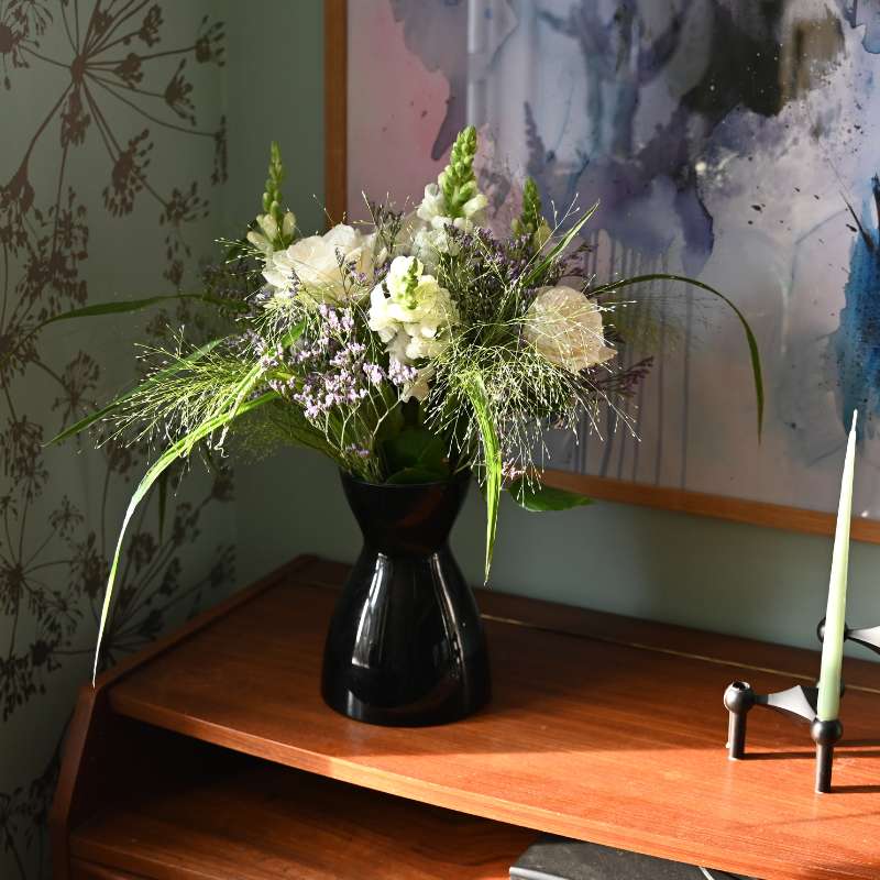 Smuk buket i hvid og blå står i en sort vase på en træhylde.