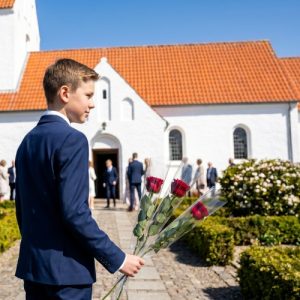 Konfirmand står med tre, enkeltindpakkede røde roser i hånden foran en hvid kirke.