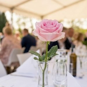 En lyserøde rose står i en glasvase på et bord til en konfirmationsfest i et udendørstelt.