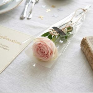 Lyserød langstilket rose indpakket i cellofan med Bloomit klistermærke på. Rosen ligger på et pænt dækket bord.