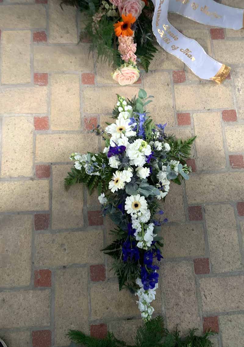 Begravelseskors lavet af blomster i blå og hvide farver. Korset ligger på et stengulv i en kirke.