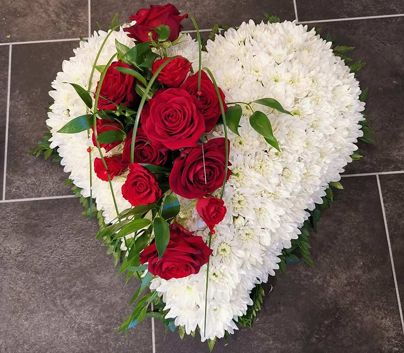 Begravelseshjerte af kun hvide blomster med en dekoration placeret ovenpå af røde roser.